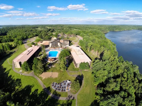 Aerial view of a resort with a pool, surrounded by green trees and a lake under a clear blue sky.