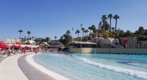 A sunny pool area with red umbrellas, palm trees, and a wave pool, surrounded by lounge chairs and a clear blue sky.