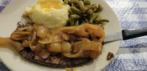 A plate with a grilled steak topped with onions, mashed potatoes, green beans, and a sunny-side-up egg.