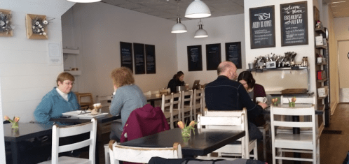 A cozy café interior with patrons at tables, flowers on each table, and a menu board on the wall.