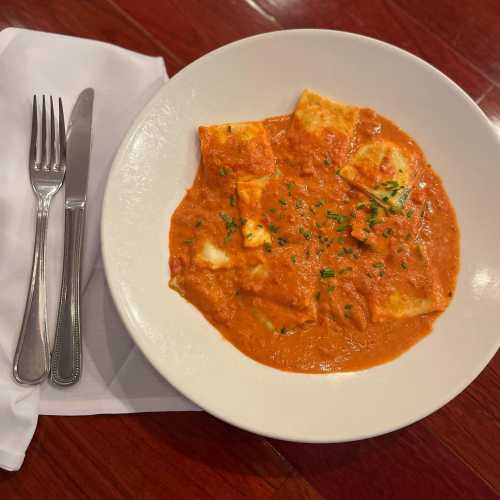 A plate of ravioli in a creamy tomato sauce, garnished with herbs, next to a knife and fork on a white napkin.