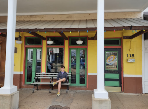 A young boy sits on a bench in front of a colorful building with green doors and festive lights.