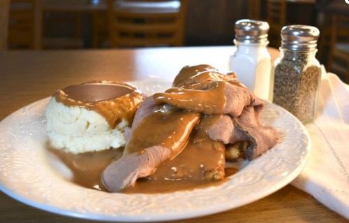 A plate of roast beef with gravy, served alongside mashed potatoes and gravy, with salt and pepper shakers in the background.