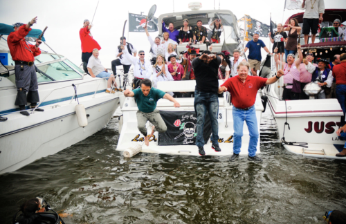 A lively group of people on boats celebrating, with some jumping into the water amidst festive decorations and flags.