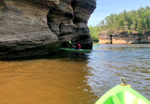 A kayaker navigates near a rocky cliff in a calm river surrounded by trees.