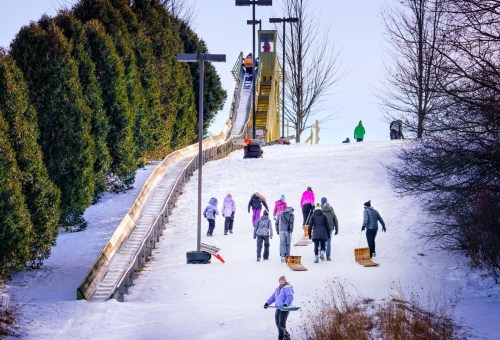 People walking up a snowy hill with sleds, heading towards a large sledding slide in a winter landscape.