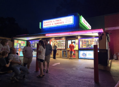 A brightly lit burger and fries stand at night, with people ordering and enjoying food outside. Neon signs glow.