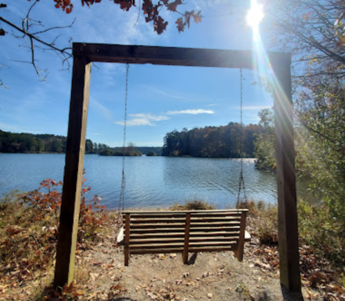 A wooden swing overlooks a serene lake surrounded by autumn foliage under a clear blue sky.