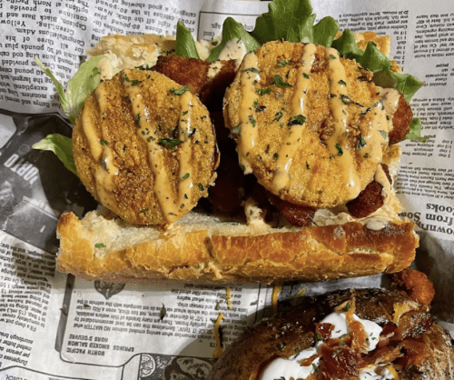 A sandwich with crispy fried green tomatoes, lettuce, and sauce on a toasted bun, served on newspaper.