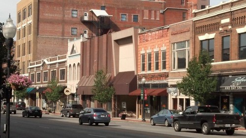 A street view of a downtown area featuring brick buildings, shops, and parked cars under a clear sky.