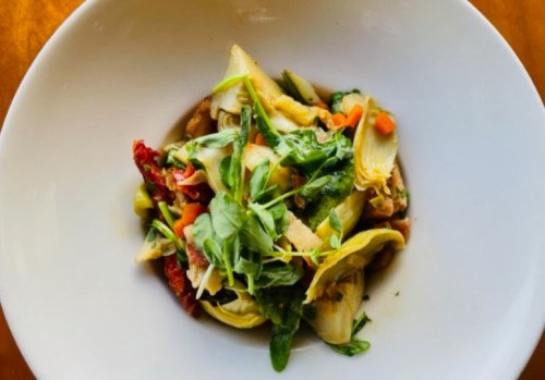 A bowl of pasta with artichokes, sun-dried tomatoes, and fresh greens, garnished with herbs.