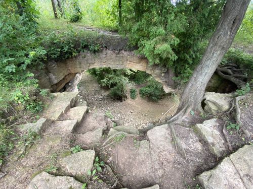 A natural stone archway surrounded by greenery, leading to a rocky, forested area below.
