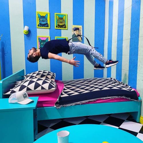 A person appears to float above a colorful bedroom, with a bed and patterned decor, creating a playful, surreal effect.