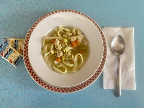 A bowl of chicken noodle soup with vegetables, accompanied by a spoon, napkin, and a pack of crackers on a blue table.