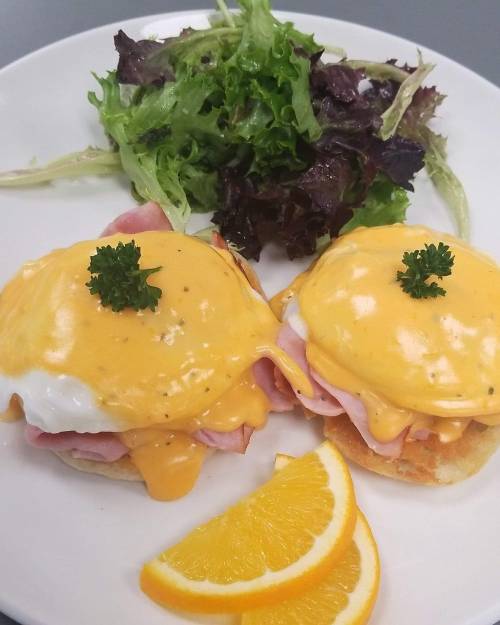 A plate of eggs Benedict with ham and cheese sauce, garnished with parsley, served with a side salad and orange slices.