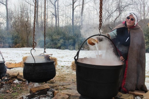 A person in historical attire pours liquid into a large pot over a fire, surrounded by snowy trees and other pots.