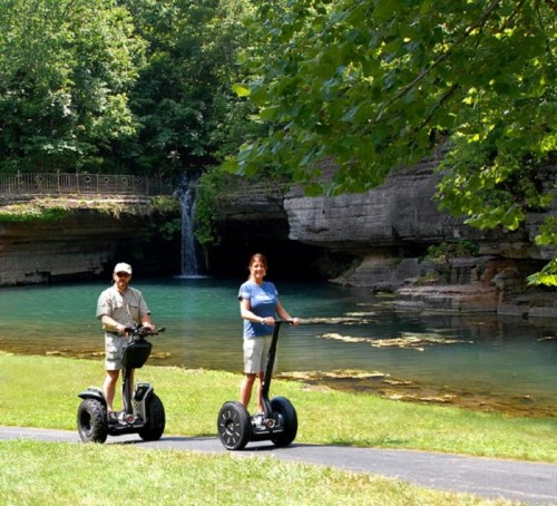 Two people riding Segways along a path near a serene, green-tinted lake and a waterfall in a lush park setting.