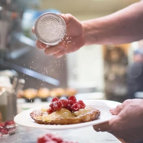 A chef sprinkles powdered sugar over a dessert topped with raspberries on a white plate.