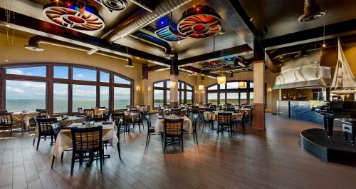A spacious restaurant with large windows overlooking the water, featuring elegant tables and modern decor.