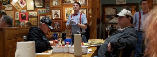 A restaurant scene with diners at tables, a waiter in a bow tie serving food, and various vintage signs on the walls.
