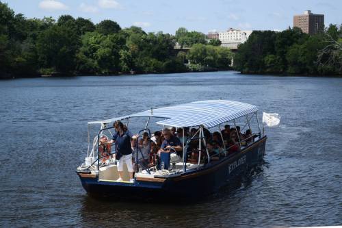 A tour boat with passengers cruises on a river surrounded by greenery and buildings under a clear blue sky.