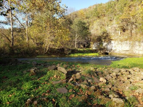 A serene river flows through a lush, green landscape with rocky banks and trees in autumn foliage.