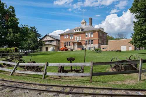 Historic building with a grassy area, surrounded by trees and old machinery, under a blue sky with fluffy clouds.