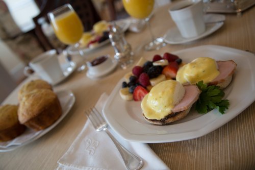 A plate of eggs Benedict with ham, garnished with fruit, muffins, and drinks on a dining table.