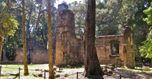 Ruins of a stone building surrounded by trees, with remnants of walls and a tower visible in a lush green setting.