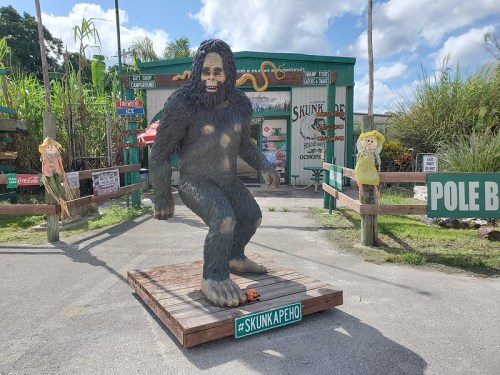 A large Bigfoot statue stands on a wooden platform outside a quirky shop, surrounded by signs and decorations.