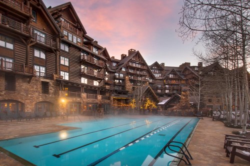 A luxurious lodge with a misty pool area, surrounded by rustic architecture and trees at sunset.