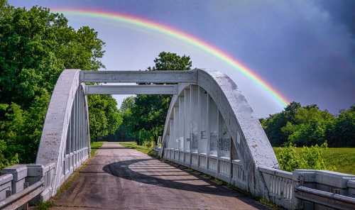 A white arch bridge spans a road, with a vibrant rainbow arcing over lush green trees in the background.