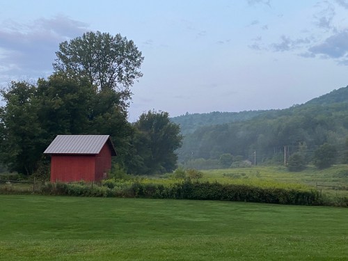 A red barn stands in a green field, surrounded by trees and rolling hills under a cloudy sky at dusk.