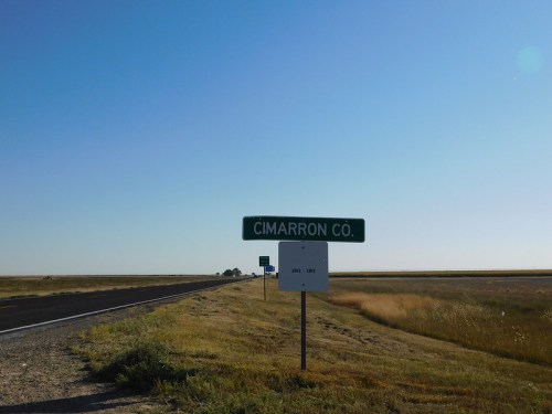 Sign for Cimarron County along a wide, open road under a clear blue sky. Fields stretch out on either side.