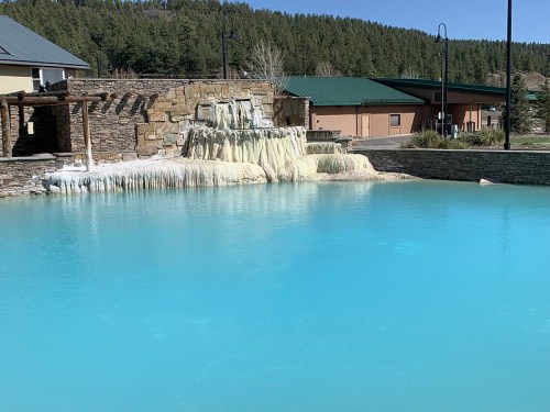 A serene blue pool with a waterfall feature, surrounded by stone structures and lush green trees in the background.