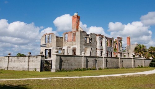 Ruins of a historic building with brick and concrete walls, surrounded by green grass and under a blue sky with clouds.