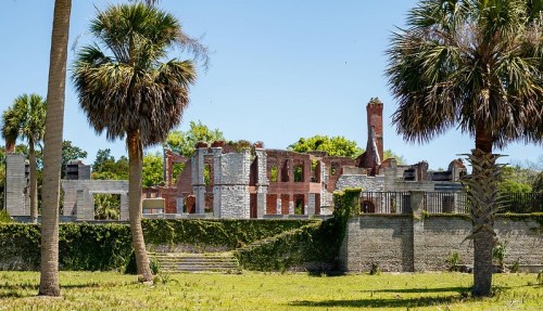 Ruins of a historic building surrounded by palm trees and greenery under a clear blue sky.