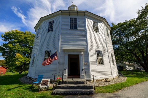 A white octagonal building with an American flag, surrounded by greenery and a blue chair on the lawn.