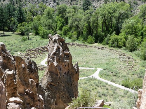 A rocky outcrop overlooks a grassy area with a winding path and a circular stone arrangement surrounded by trees.