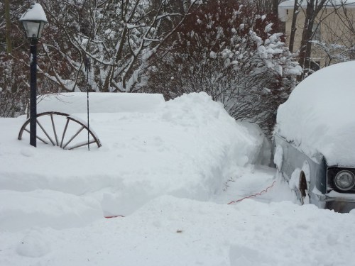 A snowy scene with a lamppost, a snow-covered car, and deep snowdrifts surrounding a path and a wheel.