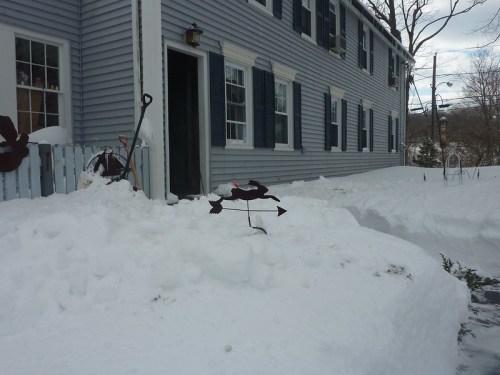 A snowy scene outside a blue house, featuring a weather vane and a snow-covered pathway.