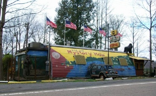 Colorful building with "Mystery Hole" sign, surrounded by trees and American flags, featuring a vintage car in front.