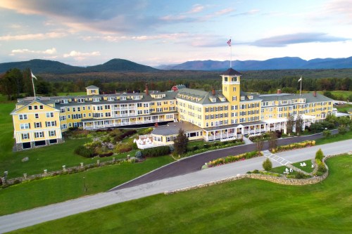 A large yellow hotel with a clock tower, surrounded by green lawns and mountains under a blue sky.