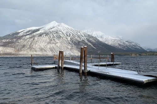 A snowy mountain rises behind a wooden dock extending into a calm lake, with overcast skies above.