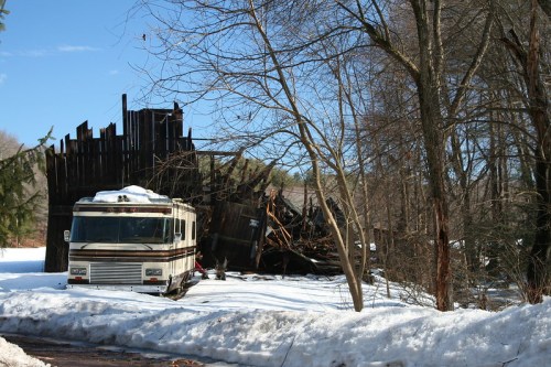 An old RV parked near a collapsed wooden structure, surrounded by snow and bare trees under a clear blue sky.