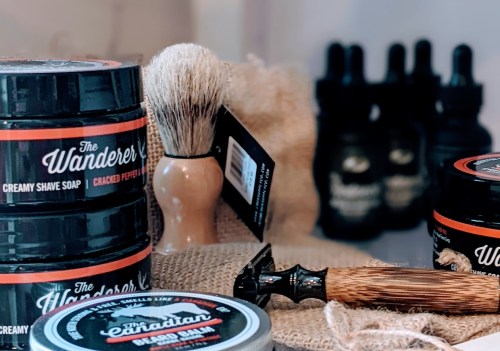 A collection of grooming products including shave soap, beard balm, a shaving brush, and a razor on a textured surface.