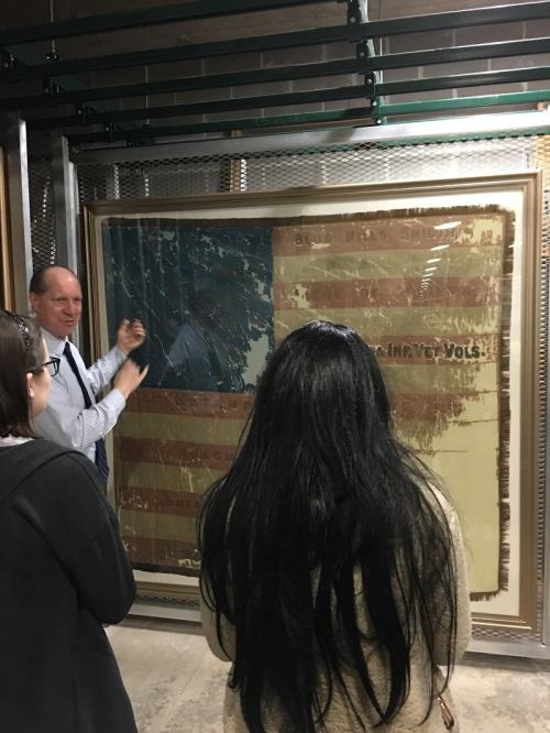 A guide explains a large, worn American flag to an audience in a museum setting.