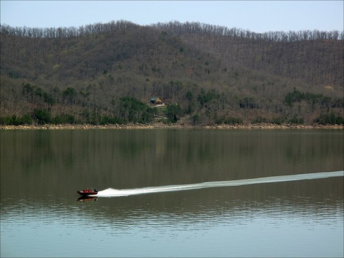 A boat speeds across a calm lake, with wooded hills and a cabin visible in the background.