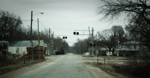A quiet, rural road with railroad crossing signals and sparse buildings on either side, under a cloudy sky.