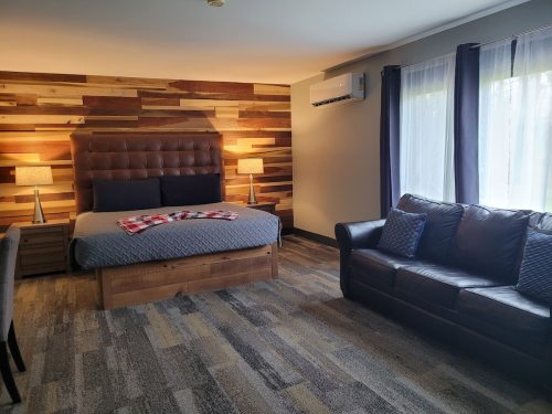 Cozy bedroom with a wooden accent wall, a bed, two lamps, and a leather sofa. Natural light from large windows.